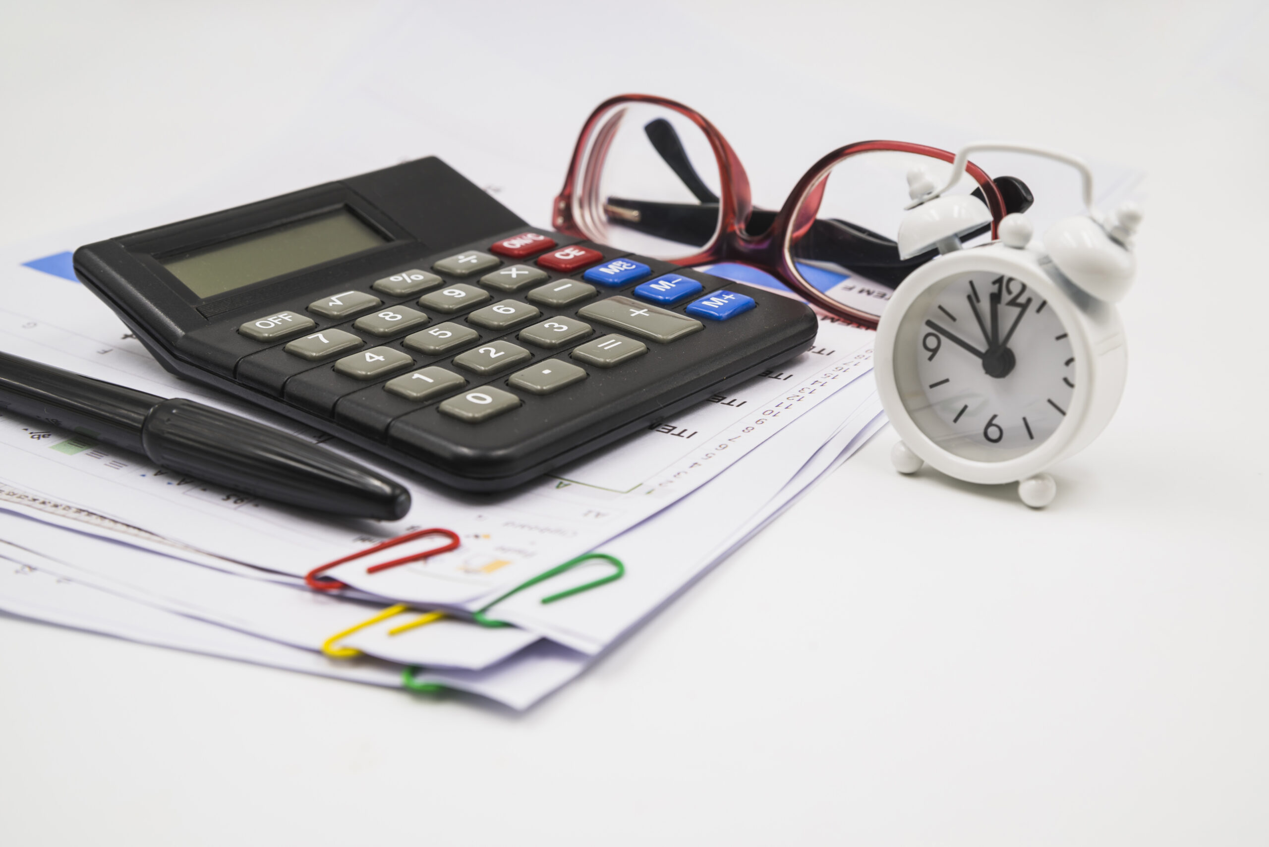 Black calculator with a pen, red-framed glasses, a white alarm clock, and stacked papers held by colorful paperclips on a desk.