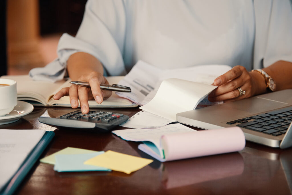 Person uses a calculator while reviewing documents at a desk with a laptop, coffee cup, and sticky notes nearby.