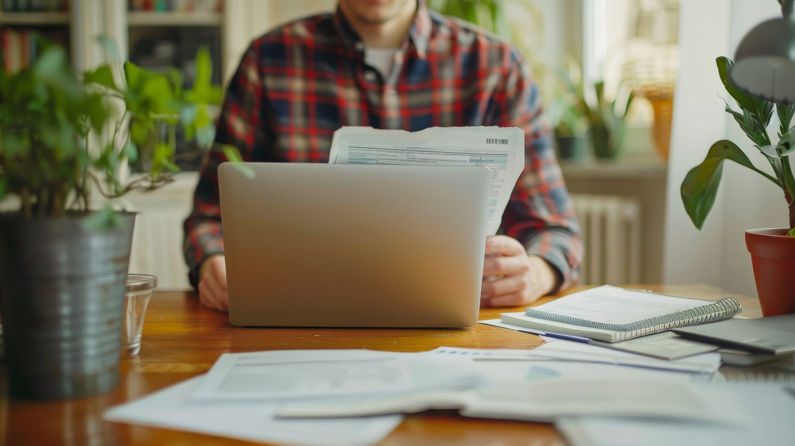 Person in a plaid shirt sits at a wooden desk with an open laptop and scattered papers, plants in view nearby.