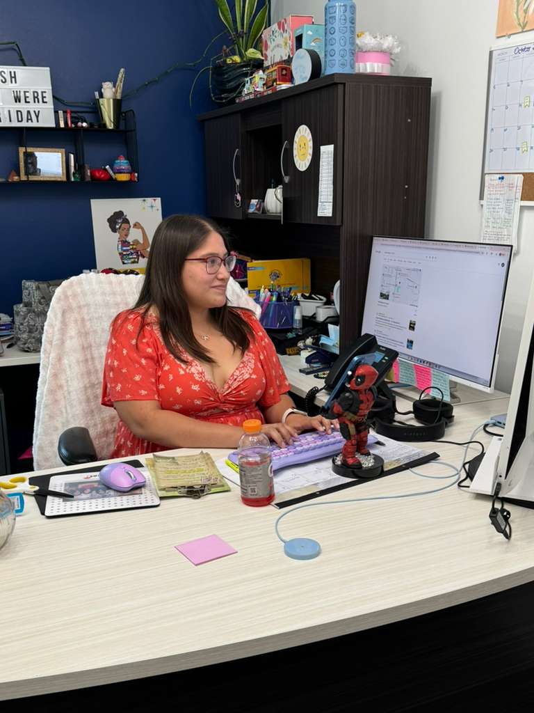 Woman in a red dress sits at a desk typing on a computer in a bright office.